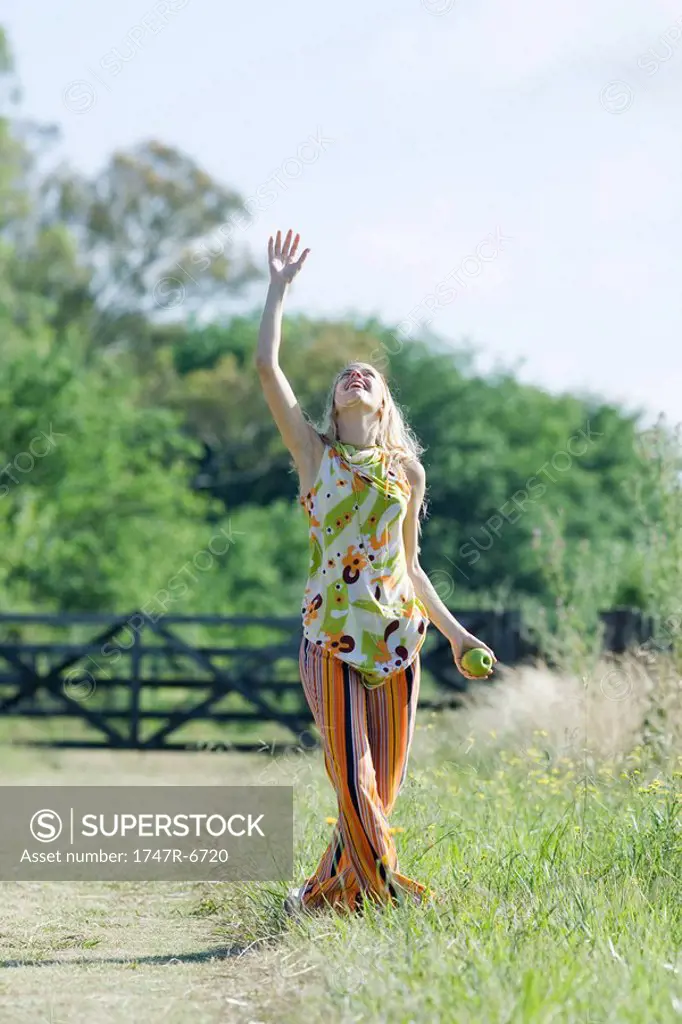 Young woman standing in rural field with arm raised, looking up, apple in hand
