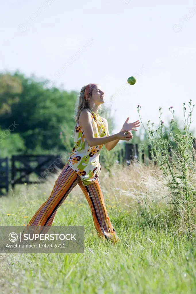 Young woman standing in field with legs apart, juggling apples