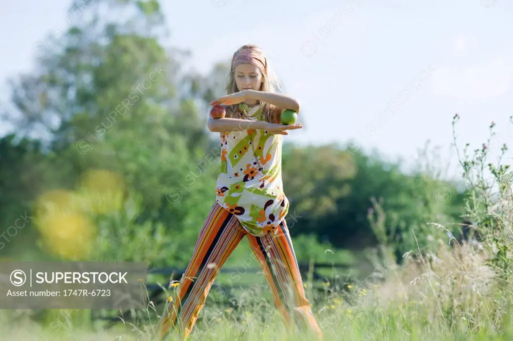Young woman standing in field, balancing apples on elbows