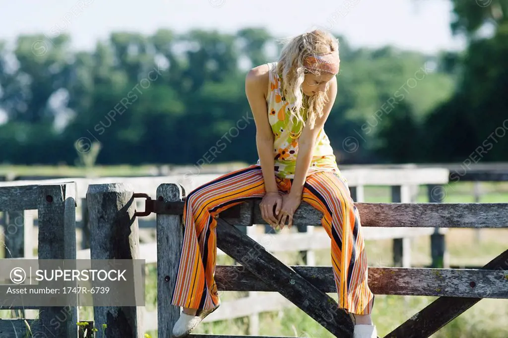 Young woman sitting on top of rural fence, looking down