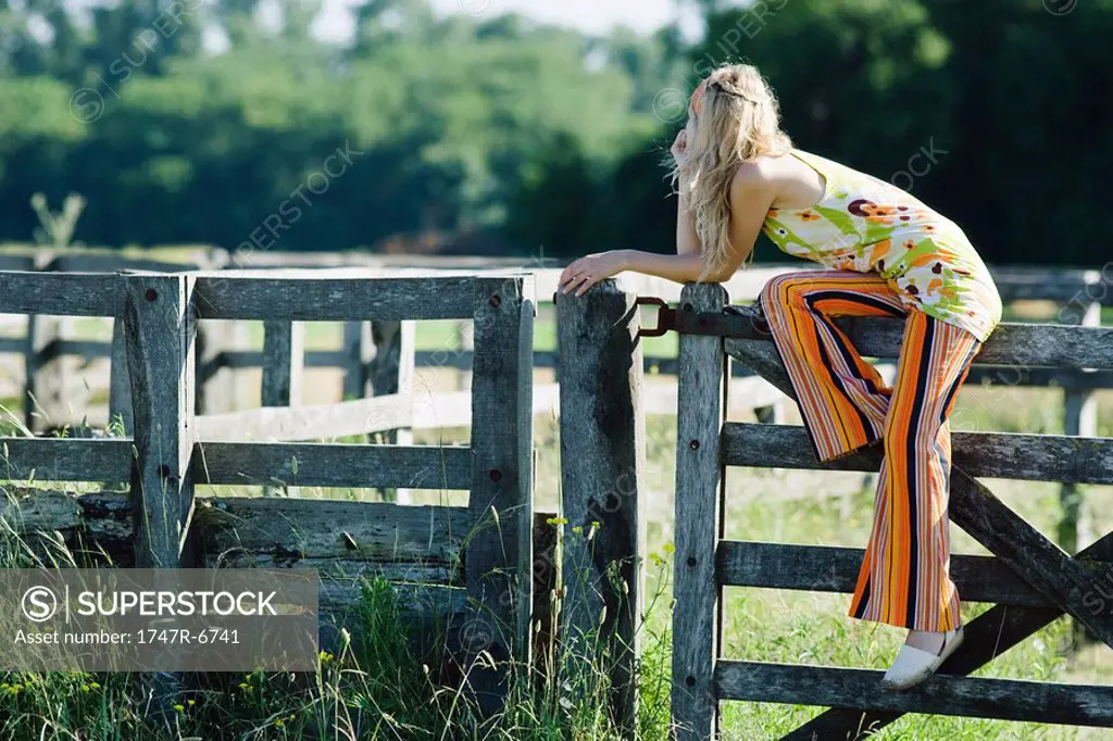 Young woman sitting on top of wooden fence, leaning on elbow, looking away