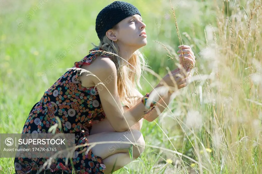Young woman crouching in field, touching tall grass and looking up