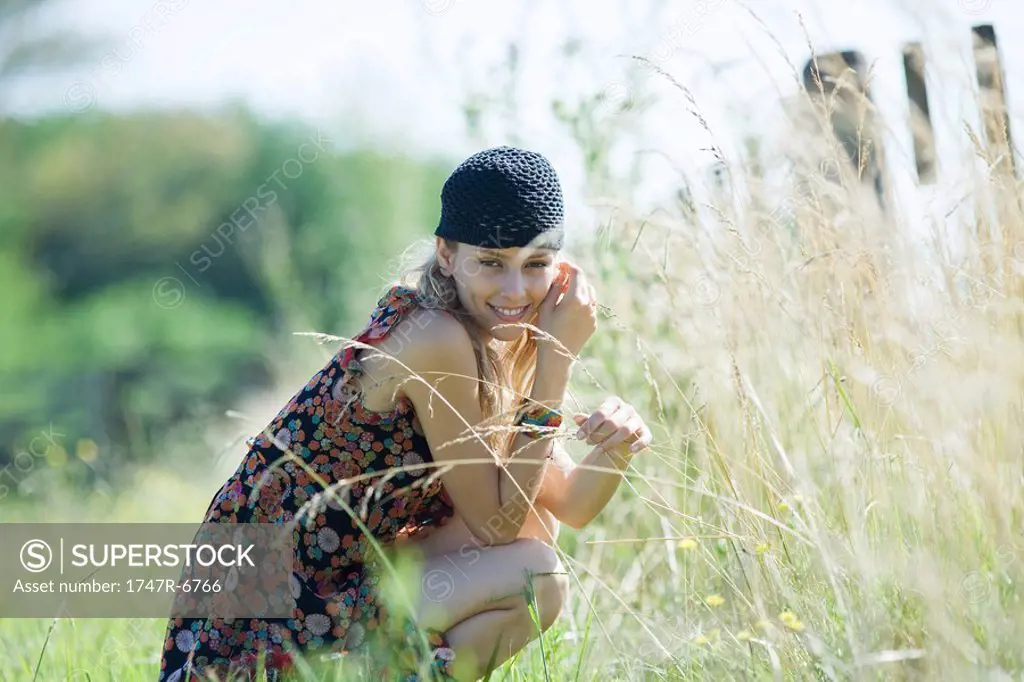Young woman crouching in field, touching tall grass and smiling at camera