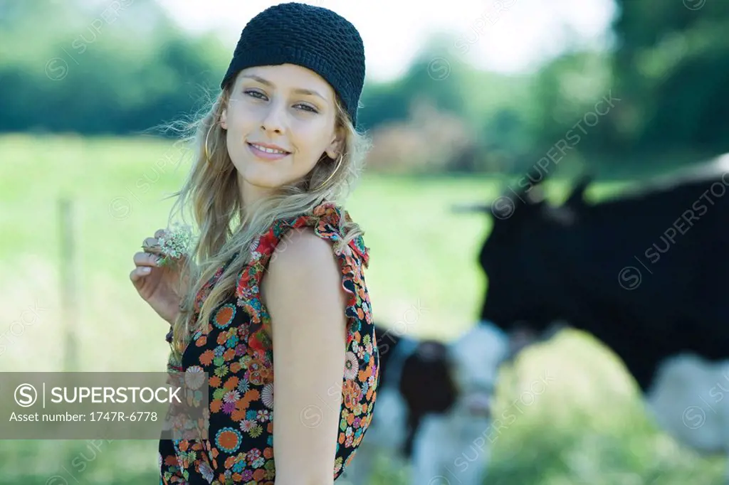 Young woman holding flower in rural setting, smiling at camera, cows in background
