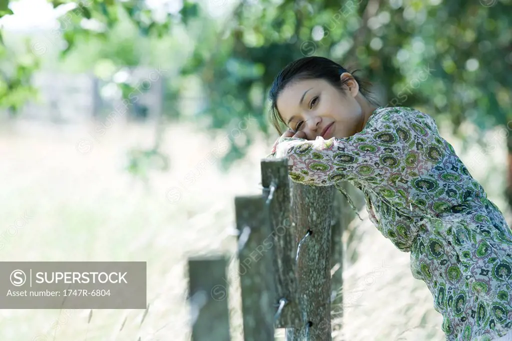 Young woman leaning against rural fence, head resting on arms, smiling at camera