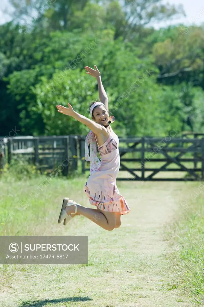 Young woman jumping with arms raised in rural setting, smiling at camera