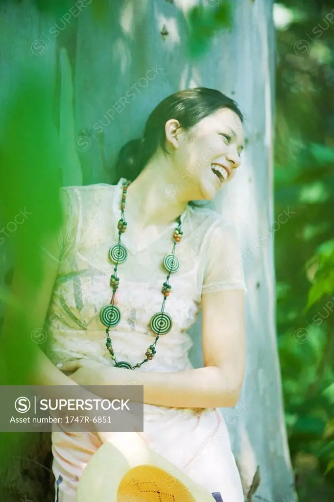 Young woman leaning against tree trunk, laughing, looking away