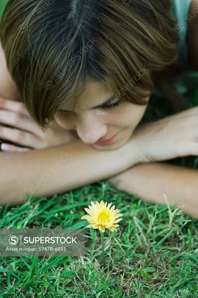 Young woman lying on grass looking at flower, head resting on arms, cropped view