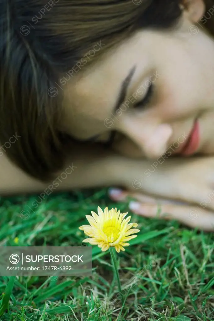 Young woman lying on grass with head resting on arms, eyes closed, flower in foreground, cropped view