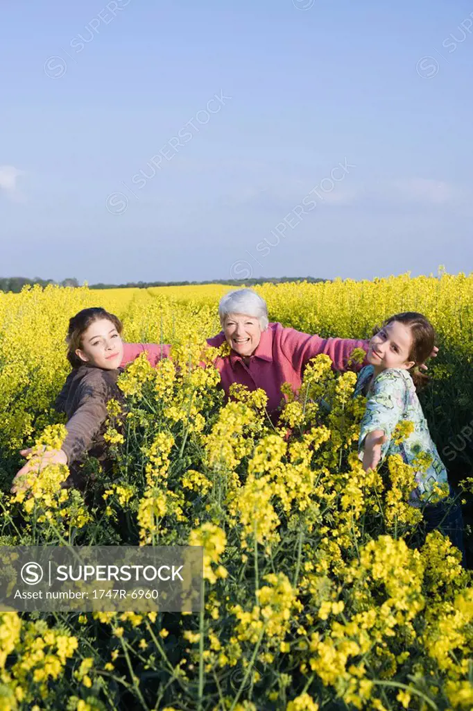 Senior woman and two granddaughters standing in field of canola in bloom, arms out toward camera