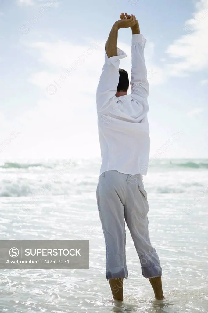 Young man standing in surf, stretching, rear view