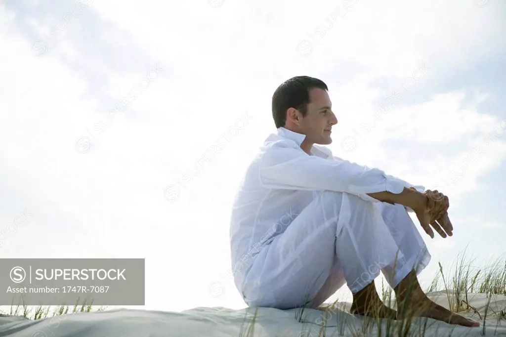 Young man sitting on dune, smiling, full length