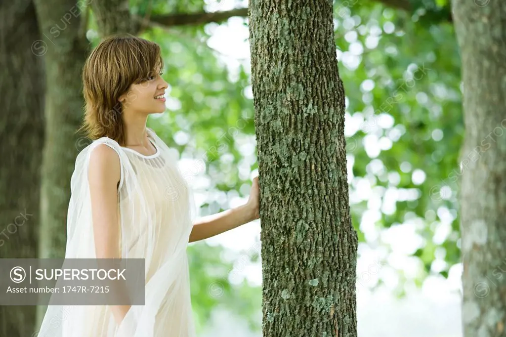 Young woman standing in woods, touching tree trunk, side view
