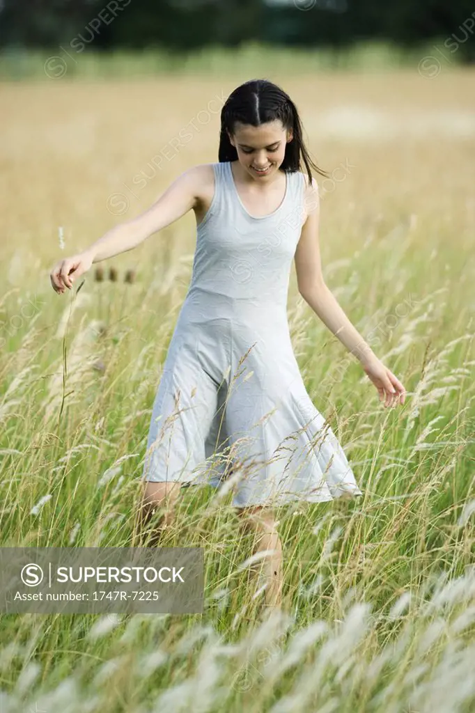 Teenage girl walking in field of tall grass with arms out, looking down