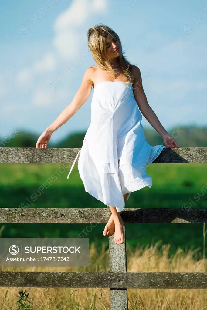 Young woman in dress sitting on rural fence with arms out, eyes closed