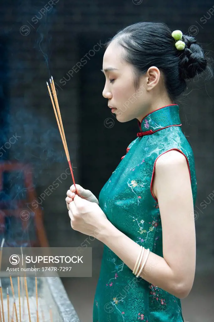 Young woman wearing traditional Chinese clothing, holding up incense, side view