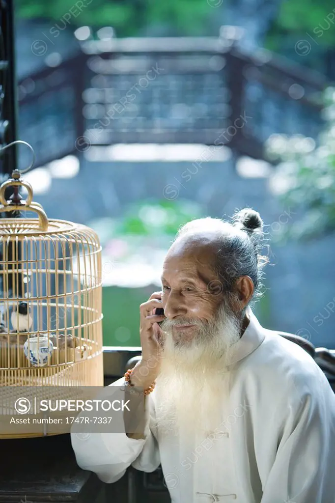 Elderly man wearing traditional Chinese clothing, using cell phone, next to bird cage
