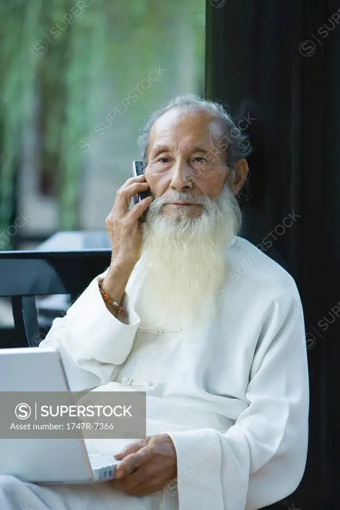 Elderly man wearing traditional Chinese clothing, using laptop and cell phone