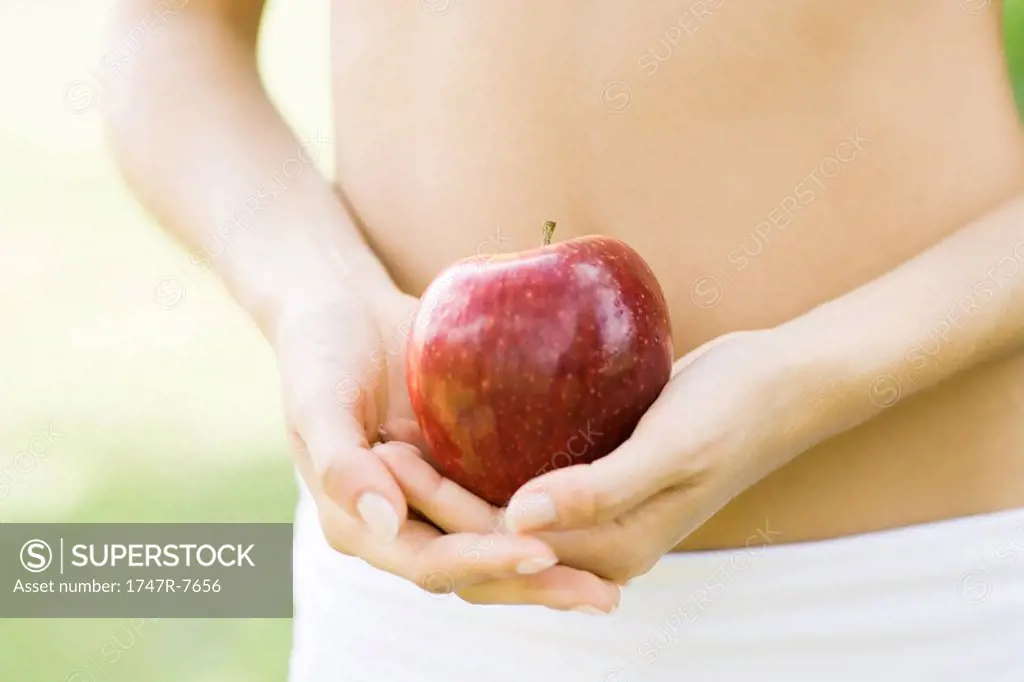 Woman holding apple in front of bare abdomen, close-up of mid section