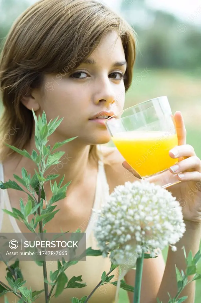 Young woman drinking glass of orange juice, plants in foreground, close-up