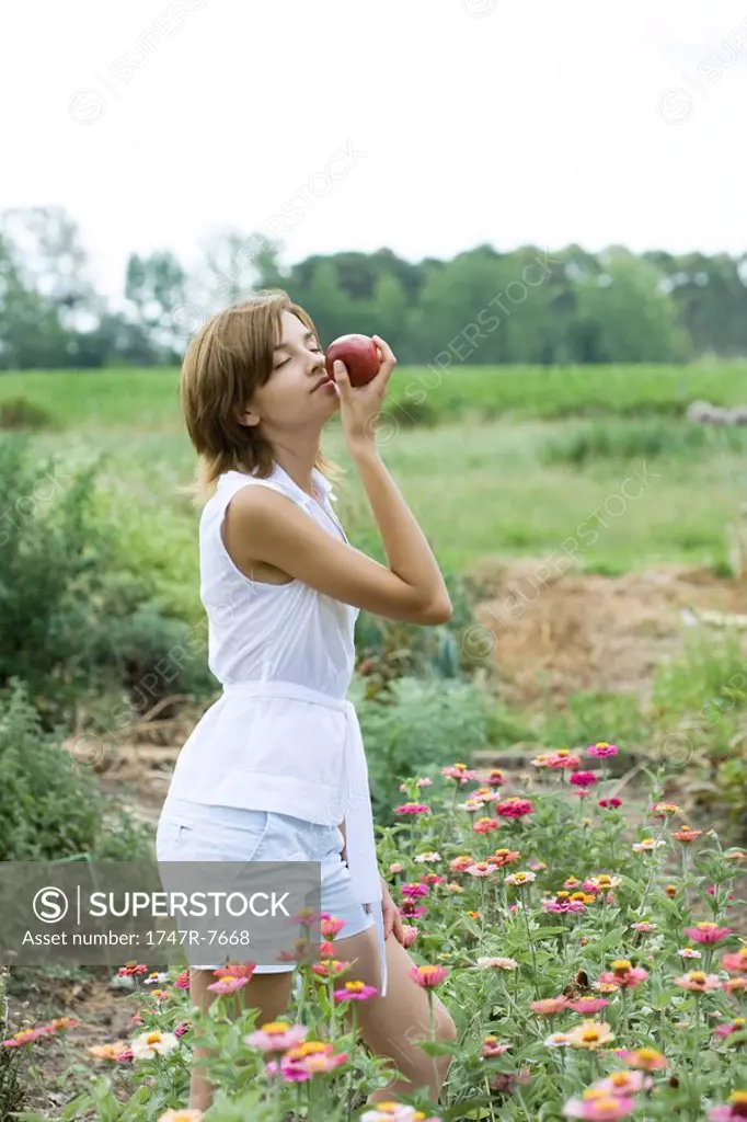Young woman standing in flower garden, smelling apple, eyes closed