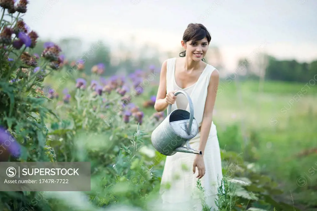 Young woman watering plants in garden with watering can, smiling at camera