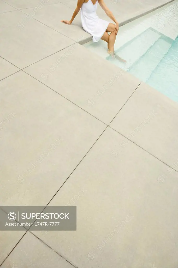 Woman sitting on edge of pool, feet in water, high angle view, cropped