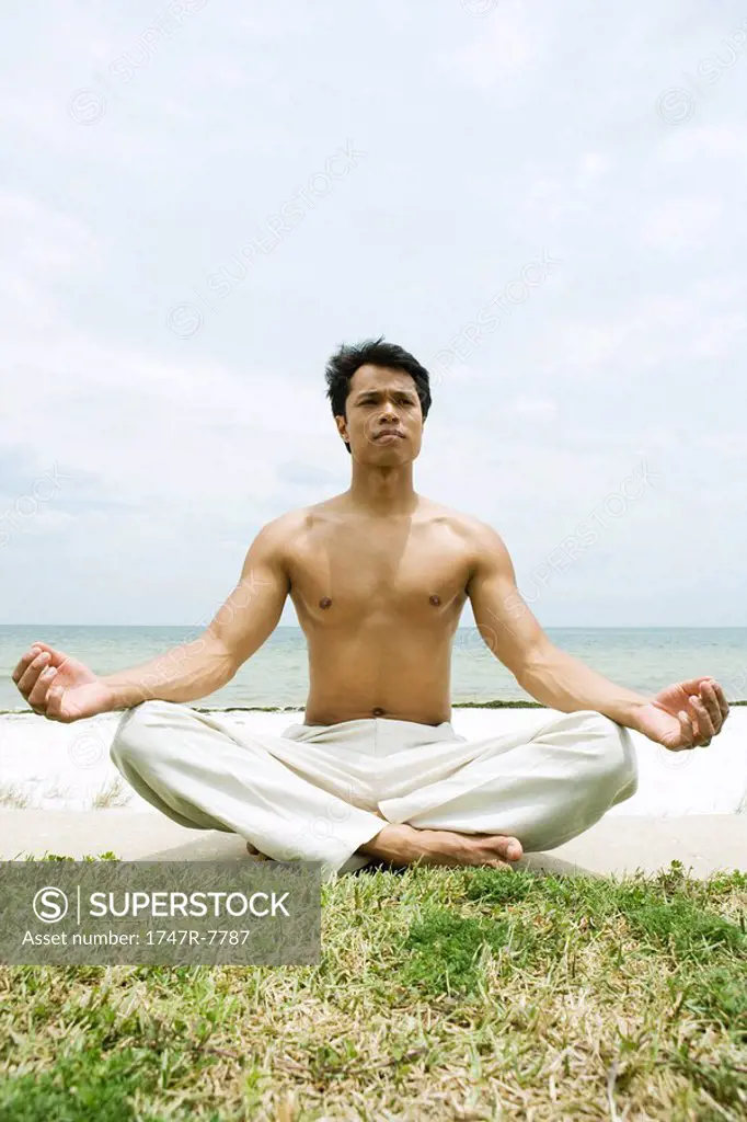 Barechested man sitting in lotus position, ocean in background, full length