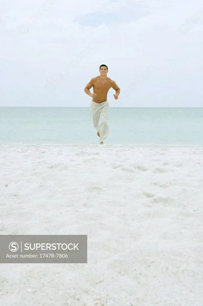 Man running on beach, facing camera, ocean in background, full length