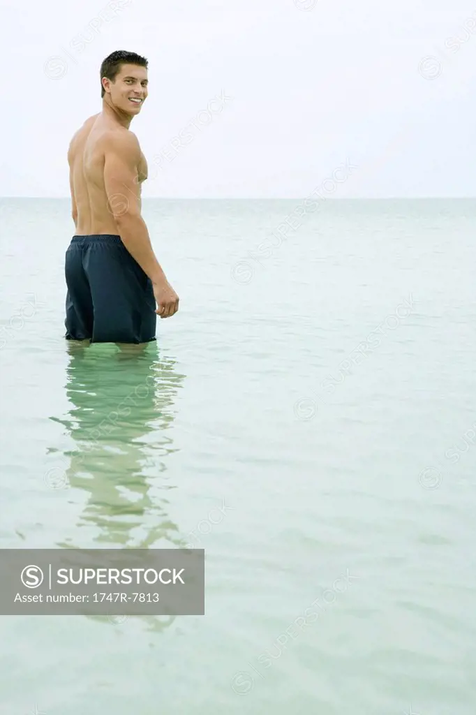 Man standing knee deep in ocean, smiling over shoulder at camera