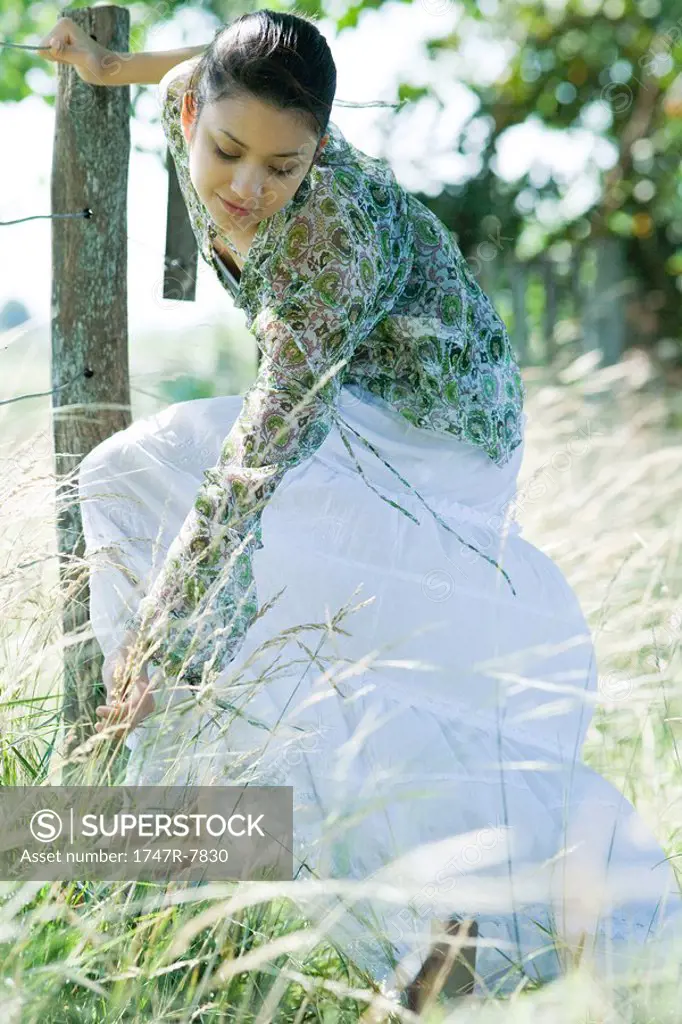 Young woman holding onto fence, bending over to pick sprig of grass