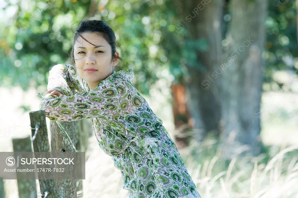 Young woman leaning against fence in rural setting, looking at camera