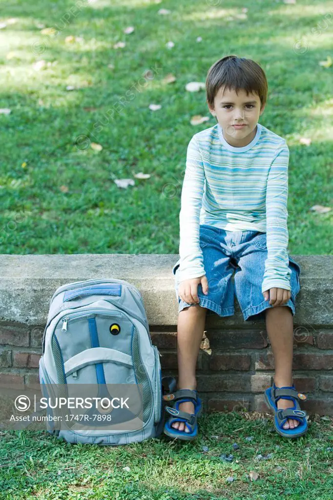 Boy sitting on low wall, backpack by side, looking at camera