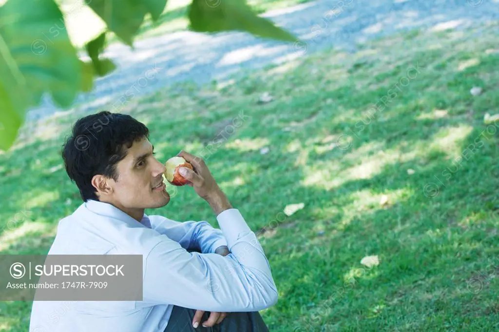 Businessman sitting on ground in park, eating apple