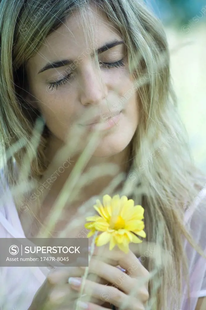 Woman holding flower, eyes closed, blurred vegetation in foreground