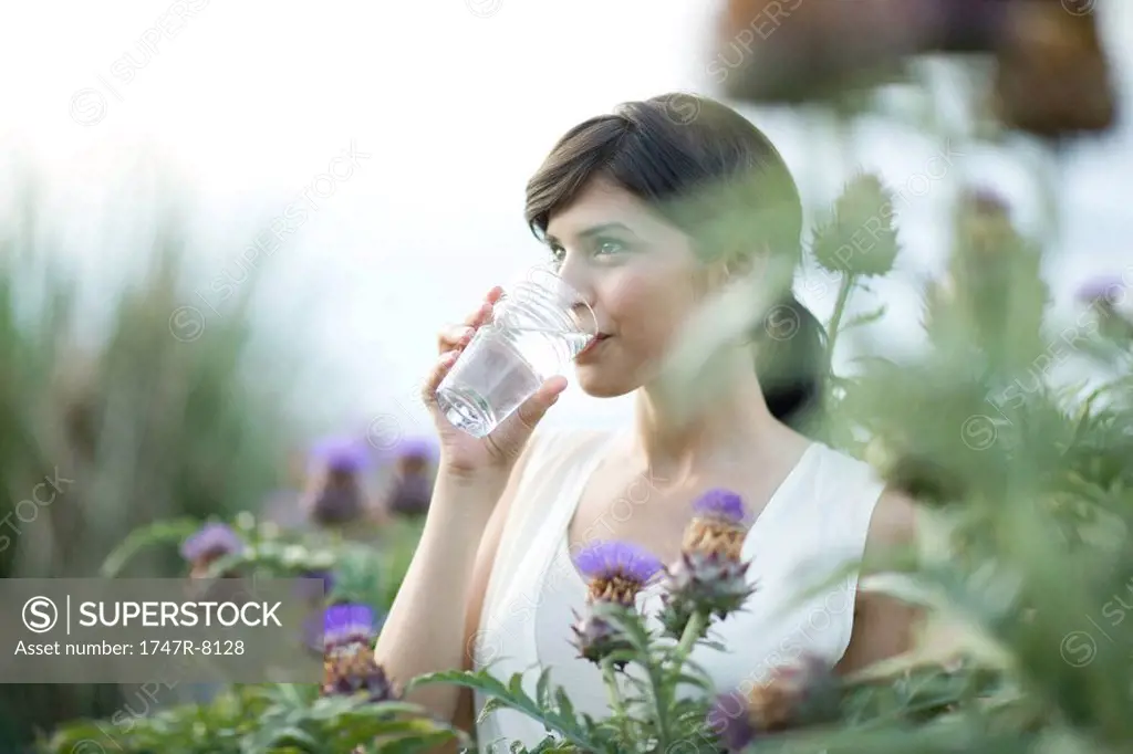 Young woman drinking glass of water outdoors, surrounded by thistle flowers