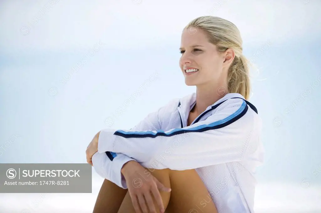 Young woman in active wear, sitting on beach, knees up, smiling
