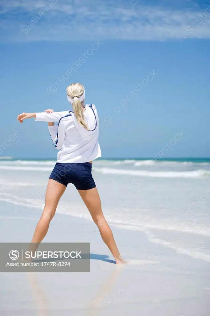 Young woman on beach, stretching, rear view