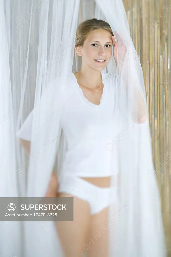 Young woman standing in underwear and tee-shirt, looking through translucent curtains, smiling at camera