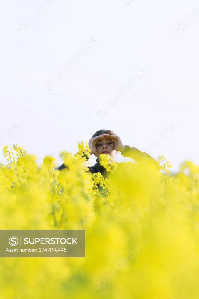 Girl standing in field of canola in bloom, shading eyes, looking at camera