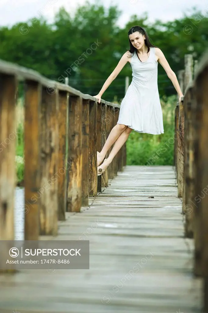 Teen girl lifting self up on railing of wooden bridge, smiling at camera