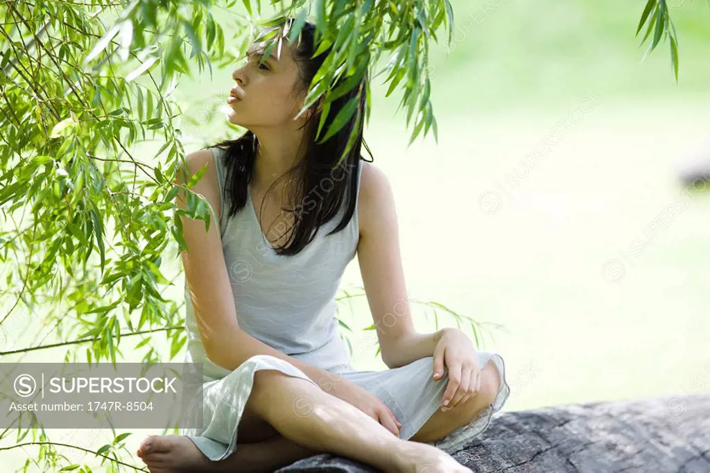 Teen girl sitting on log, under tree, looking away