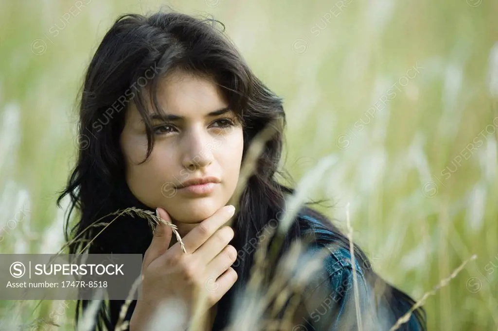 Young brunette woman in field, hand under chin, looking away, portrait