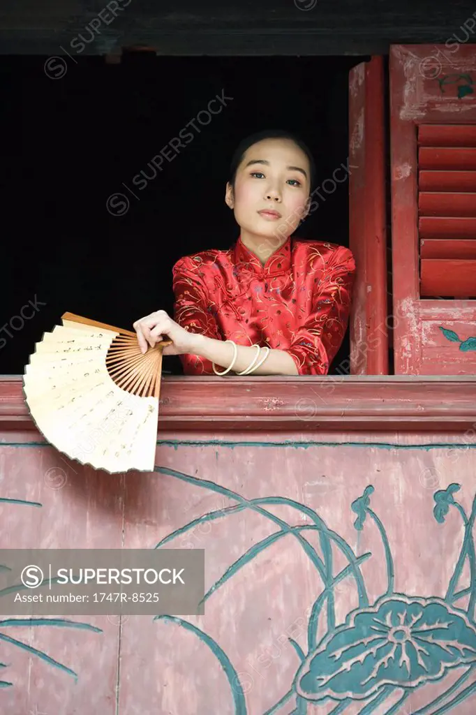 Young woman dressed in traditional Chinese clothing, leaning out of window sill, holding fan