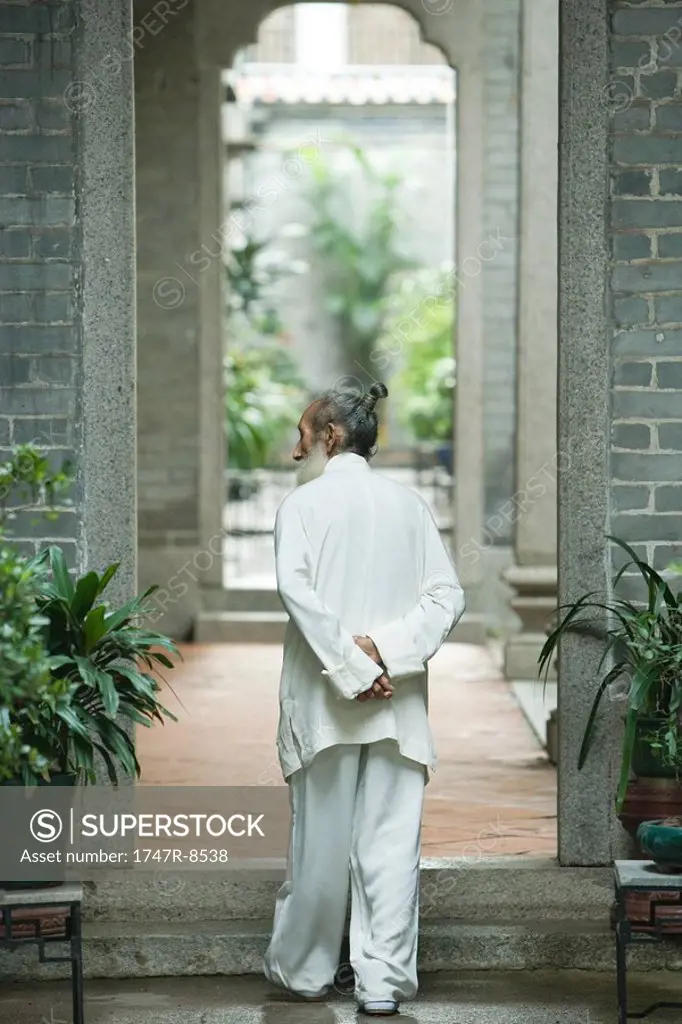 Elderly man in traditional Chinese clothing, walking through archway with hands behind back