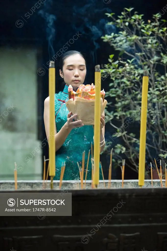 Young woman dressed in traditional Chinese clothing holding up religious offering near burning incense