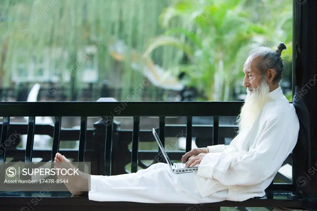Elderly man in traditional Chinese clothing, barefoot, using laptop