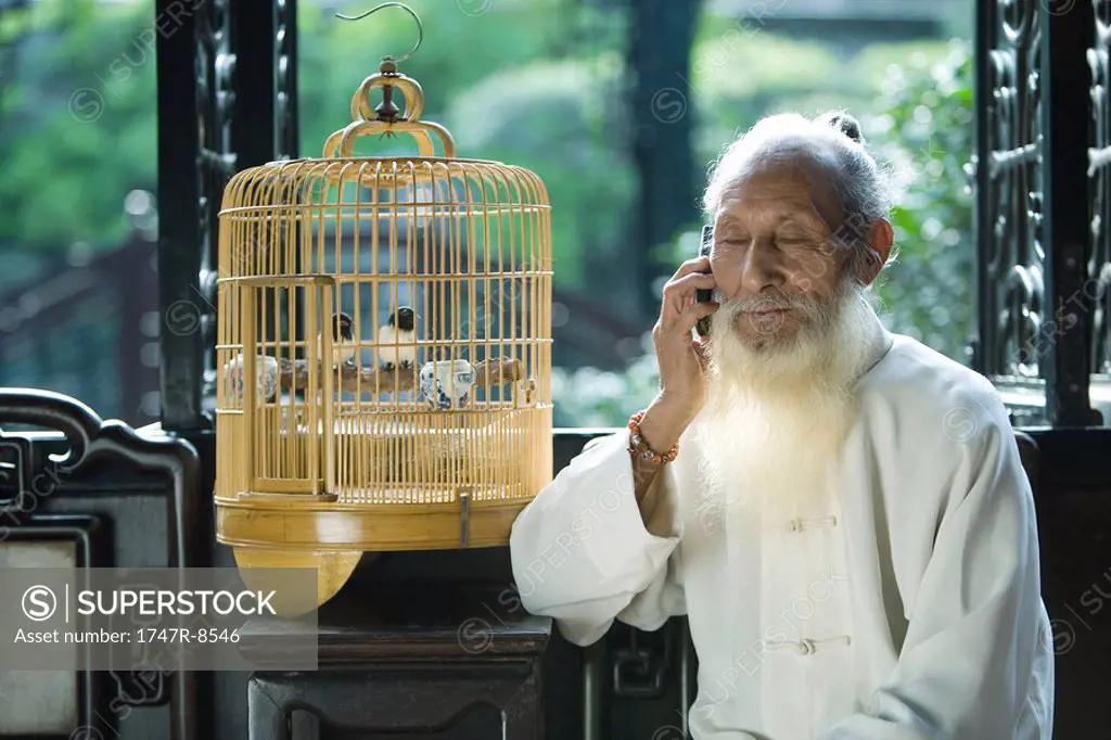 Elderly man in traditional Chinese clothing holding cell phone to ear, sitting next to bird cage