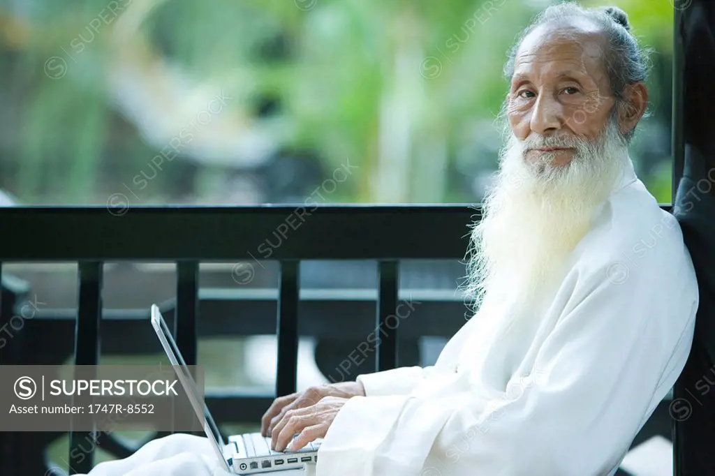 Elderly man in traditional Chinese clothing using laptop computer, looking at camera