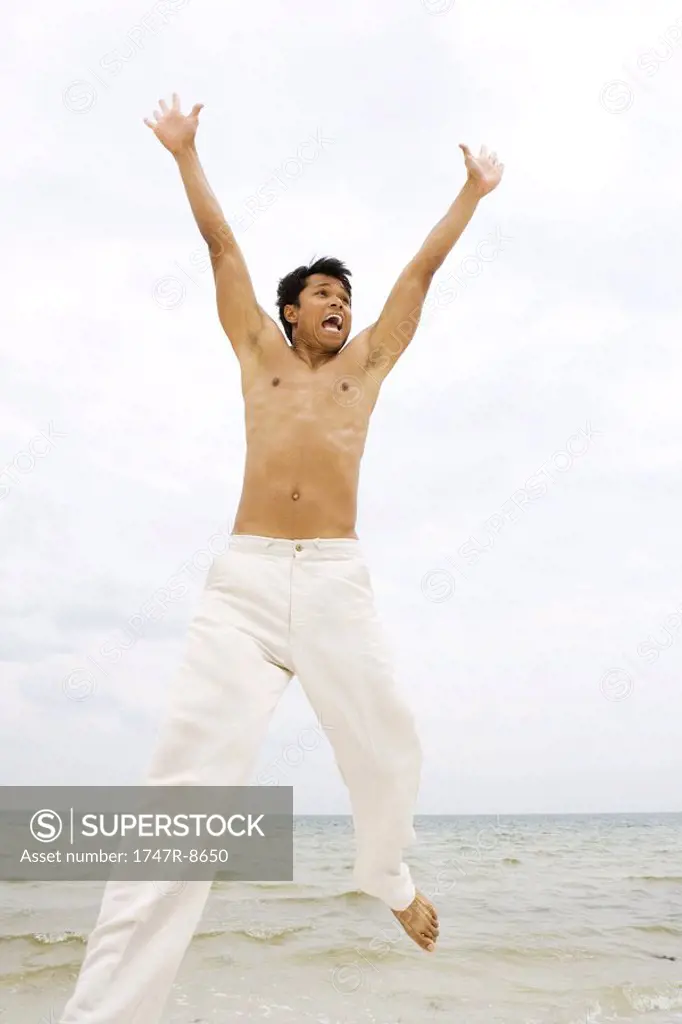 Man jumping in the air at the beach, arms raised, looking away